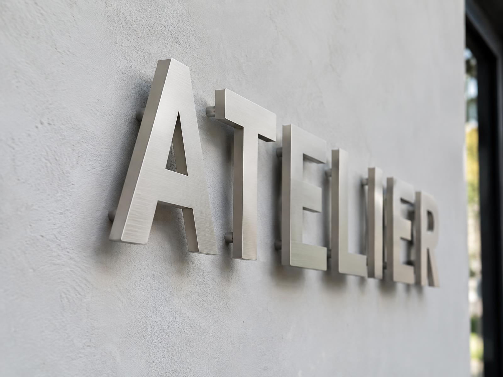 Geborsteld zilveren 3D-letters 'ATELIER' zwevend voor een stucmuur op verborgen afstandhouders